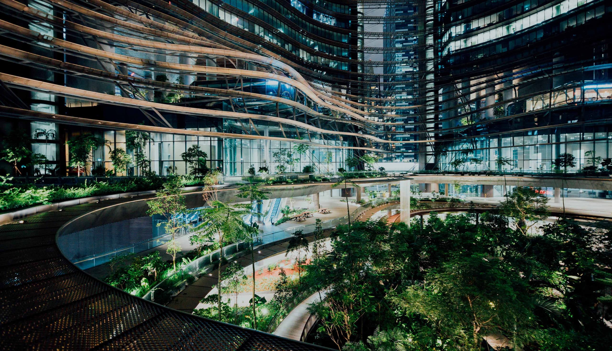 Espacio interior moderno con jardines, pasarelas curvas y edificios de vidrio iluminados por la noche.